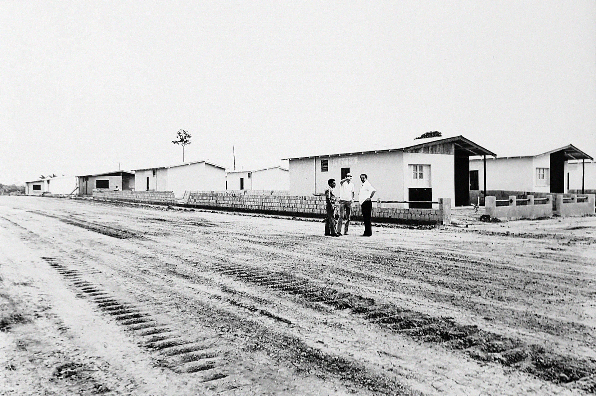 Fotografia de arquivo em preto e branco: obra da Construtora Flávio Espírito Santo em Manaus, com três homens numa rua de terra diante de casas recém-construídas.