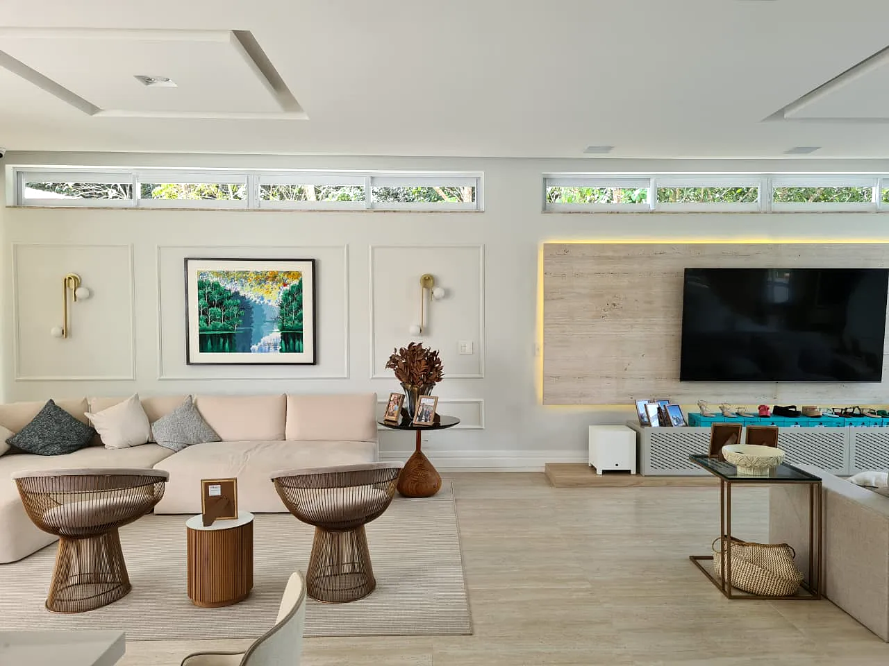 Living com sofá claro, poltronas em arame, painel em travertino e quadro com paisagem amazônica.